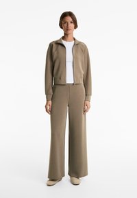 Tuta beige a costine con giacca con zip e pantaloni a gamba larga, con una t-shirt bianca sotto. Tessuto testurizzato, design aderente.