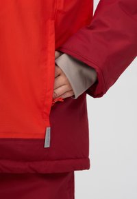 Veste rouge et orange avec une poche latérale zippée, poignet gris et étiquette de logo. Présente des couleurs contrastées et un tissu lisse et léger.