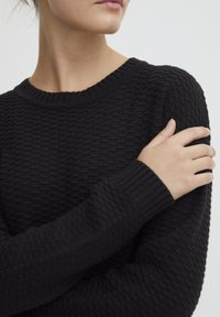 Maglione nero lavorato a maglia con un motivo testurizzato, colletto e polsini a coste, e una vestibilità rilassata. In primo piano si vede un braccio che abbraccia la spalla.