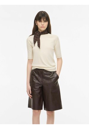 Mujer con una camiseta beige de manga corta, pantalones cortos de cuero marrón hasta la rodilla y una bufanda marrón de cuadros atada al cuello, de pie frente a un fondo blanco.