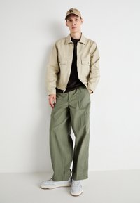 Veste zippée beige avec deux poches avant, t-shirt noir, pantalon large vert olive et baskets gris clair. Tenue décontractée, tons neutres.