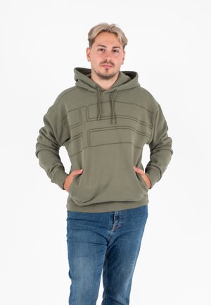 Olivengroene hoodie met een voorzak, een trekkoord kap, en een getextureerd geometrisch lijnontwerp op de borst. Draagt met blauwe jeans.