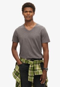 Camiseta gris de cuello en V con mangas cortas, combinada con una camisa de franela verde y a cuadros atada a la cintura, llevada con jeans oscuros.