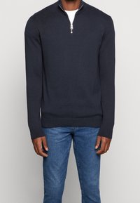 Maglione zip-up navy con maniche lunghe, realizzato in un morbido materiale knit. Presenta un colletto alto e polsini e orlo a coste. Abbinato a jeans blu.