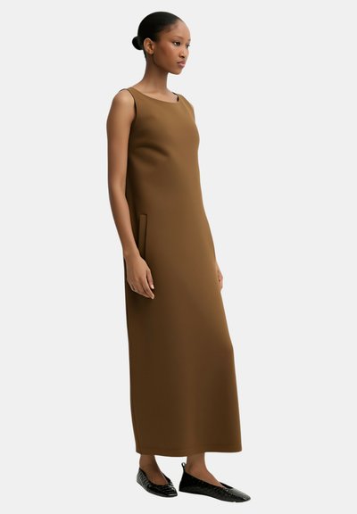 Vestito lungo marrone senza maniche con silhouette dritta, caratterizzato da una tasca laterale e una texture liscia. Indossato con scarpe nere piatte.