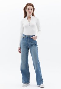 Haut blanc à manches longues avec un col à demi-fermeture éclair, associé à un jean large taille haute bleu clair. Le mannequin porte des chaussures blanches.