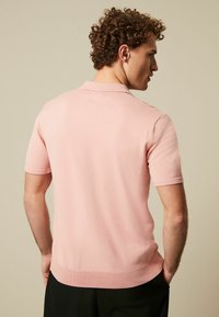 Polo de culoare roz, cu mânecă scurtă, fabricat dintr-un material tricotat moale, având un design cu guler și margine ribbed. Fără modele sau accente proeminente.