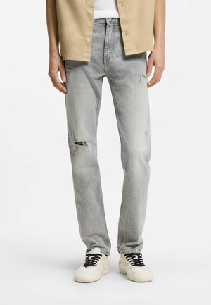 Hellgraue, abgenutzte Jeans mit Rissen am linken Knie und rechten Oberschenkel, getragen mit weißen Sneakers mit schwarzen Schnürsenkeln und beige Hemd über weißem Top.
