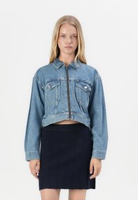 Denim cropped jacket in light blue, featuring a zip front, two chest pockets, and long sleeves. Paired with a black ribbed skirt.