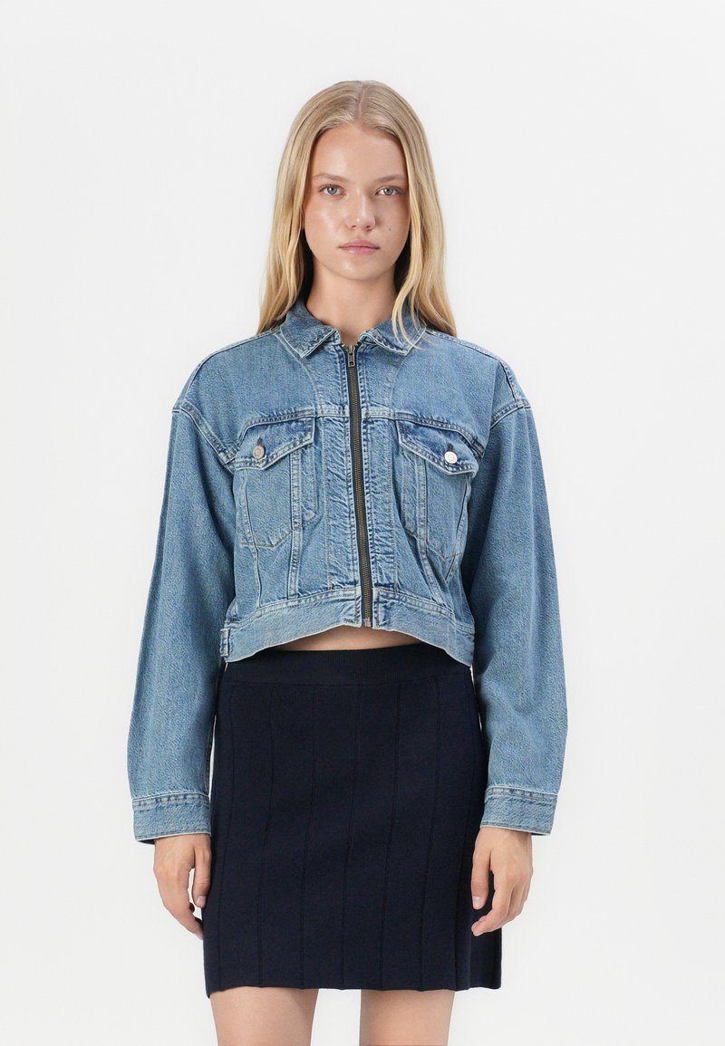 Denim cropped jacket in light blue, featuring a zip front, two chest pockets, and long sleeves. Paired with a black ribbed skirt.