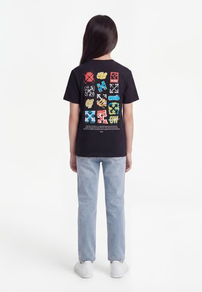 Παιδί με μακριά σκούρα μαλλιά, που στέκεται γυρισμένο με την πλάτη, φοράει μαύρο γραφικό μπλουζάκι με πολύχρωμα λογότυπα, ανοιχτό μπλε τζιν και λευκά αθλητικά παπούτσια.