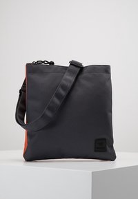 Šedá látková taška s oranžovým bokem, vybavená černým nastavitelným popruhem a obdélníkovou černou šikmou nášivkou. Hladká textura, minimalistický design.