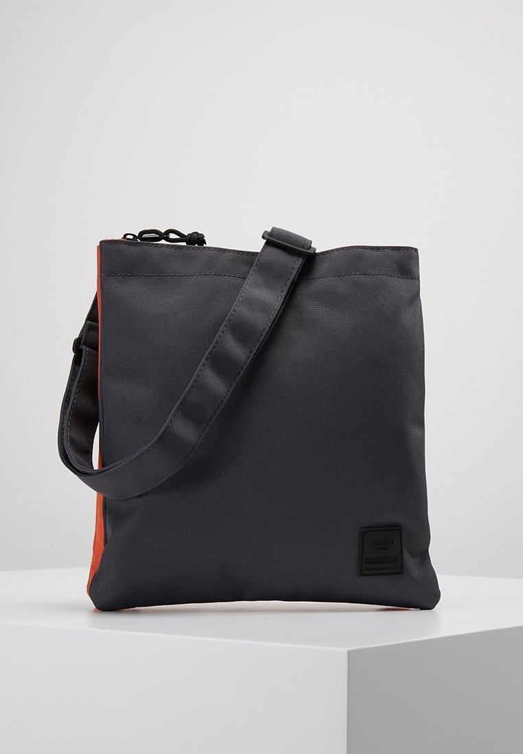 Šedá látková taška s oranžovým bokem, vybavená černým nastavitelným popruhem a obdélníkovou černou šikmou nášivkou. Hladká textura, minimalistický design.