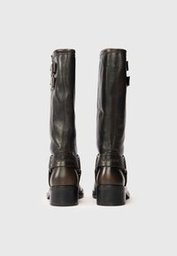 Un par de botas de cuero marrón oscuro hasta la mitad de la pantorrilla con tacones de bloque y correas con hebillas, vistas desde atrás sobre un fondo blanco.