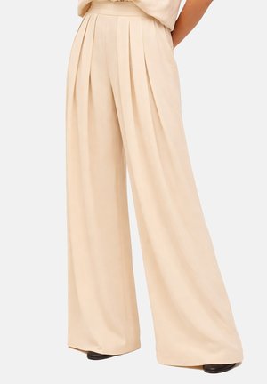 Pantalon beige taille haute à jambes larges avec plis avant, porté avec des chaussures noires sur un fond uni.