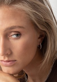 Silver hoopörhängen med små klara ädelstenar. Slät metallyta med en polerad finish, bärs på en modell med ljust blont hår.