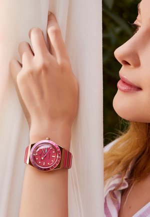 Montre en quartz rose avec un cadran rond, des accents dorés et un bracelet en caoutchouc rayé rose. Ses caractéristiques notables incluent un affichage de la date et des marqueurs luminescents.