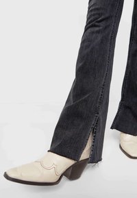 Jean en denim noir avec ourlets bruts et fentes latérales, associé à des bottines cowboy crème à talons pointus avec coutures décoratives.