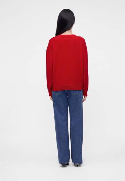 Femme aux longs cheveux noirs portant un pull rouge et un jean bleu, debout de dos sur un fond blanc uni.