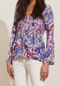 Lila blommig blus med långa trumpetärmar. Har ett smockat livstycke och visar rosa, blå och gröna blommönster på lätt tyg.