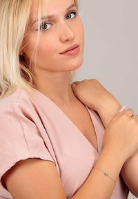 Silver bracelet with a round charm, worn on a wrist. Light pink fabric top, soft texture, V-neck design, with a neutral background.