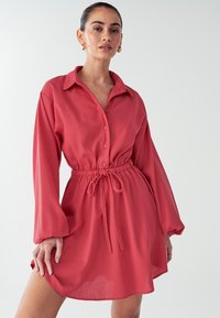 Rote, knöpfbare Hemdkleid mit langen Ärmeln, tailliertem Schnitt und lockerem Sitz. Hergestellt aus einem leichten Stoff mit glatter Textur.