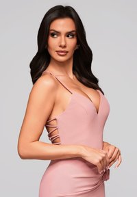 Kobieta o długich ciemnych włosach, ubrana w różową sukienkę bez rękawów z bocznymi paskami, patrząca w lewo na tle jednolitego jasnego tła.