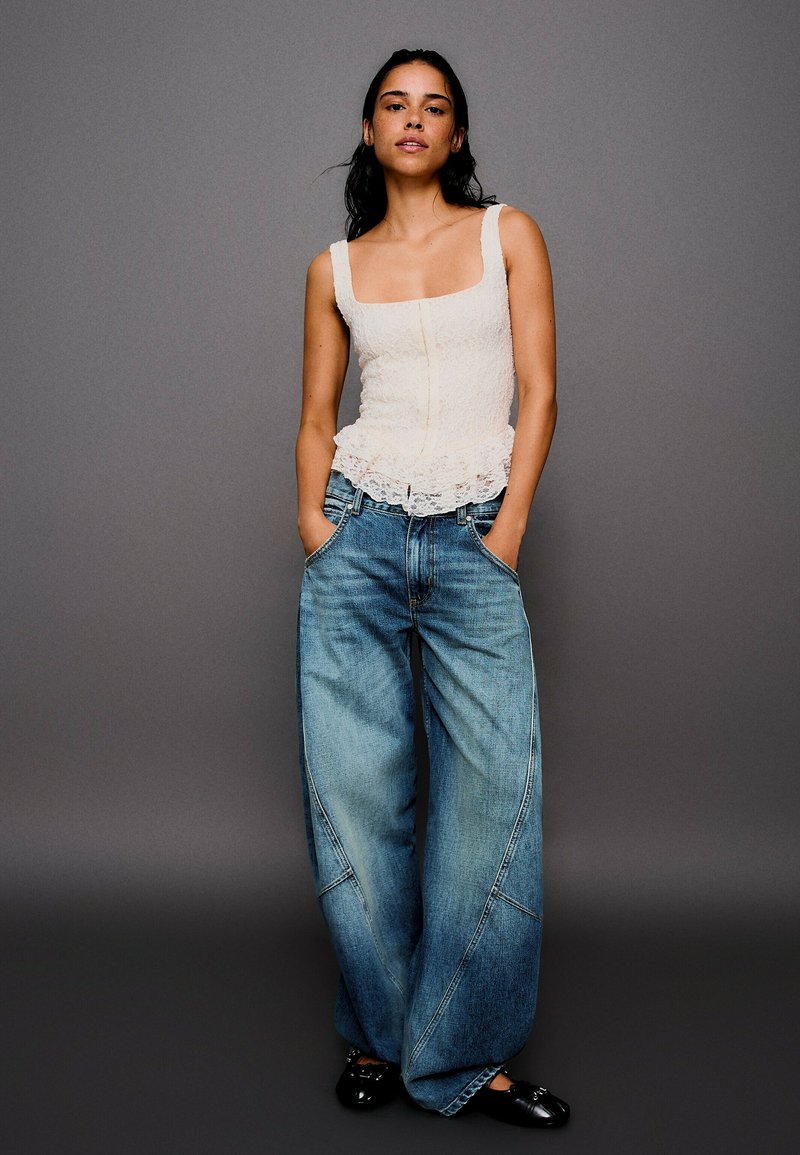 Femme aux longs cheveux foncés portant un top sans manches en dentelle blanche, un jean bleu à jambes larges, des chaussures noires, debout, les mains dans les poches, sur fond gris.