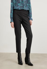 Svarta skräddarsydda byxor med rak passform, tillsammans med en långärmad topp med ett blått och grönt marmoreringmönster. Bärs med svarta hälsäkrade ankelskor.