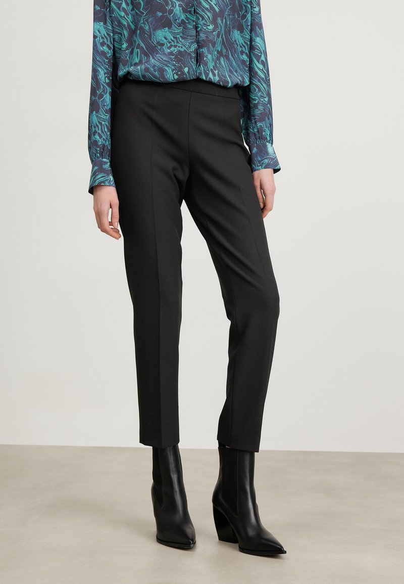 Svarta skräddarsydda byxor med rak passform, tillsammans med en långärmad topp med ett blått och grönt marmoreringmönster. Bärs med svarta hälsäkrade ankelskor.