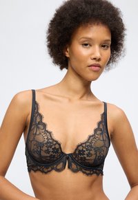Γυναίκα με σγουρά μαλλιά που φοράει μαύρο δαντελένιο σουτιέν με λουλουδένια σχέδια, ποζάροντας μπροστά σε απλό ανοιχτόχρωμο φόντο.