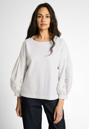 Woman with long dark hair wearing a white blouse with balloon sleeves featuring eyelet embroidery, paired with dark jeans, standing against a plain background.