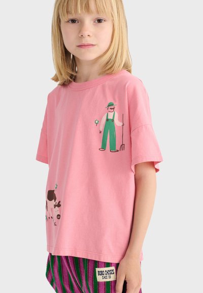 Małe dziecko o blond włosach nosi różową koszulkę z ilustracją rolnika trzymającego widły i krowy oraz paskowane spodenki.