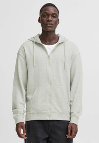 Felpa con zip di colore grigio chiaro, dotata di cappuccio regolabile con coulisse, grandi tasche frontali e polsini a coste. Tessuto morbido e vestibilità rilassata.