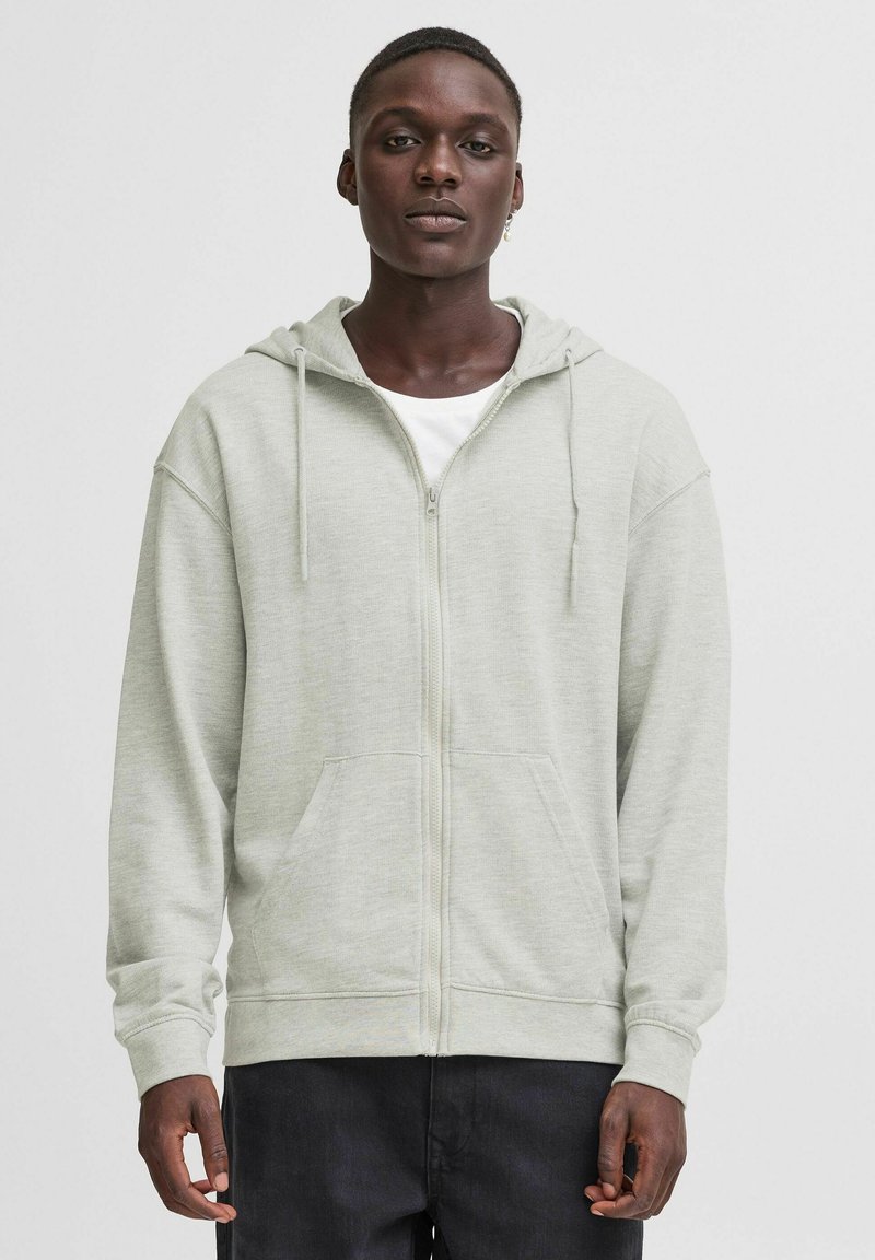Felpa con zip di colore grigio chiaro, dotata di cappuccio regolabile con coulisse, grandi tasche frontali e polsini a coste. Tessuto morbido e vestibilità rilassata.