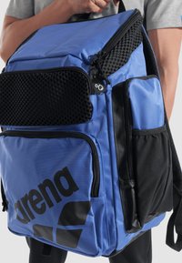 Mochila azul con un bolsillo frontal de malla texturizada, acentos negros y un logotipo negro prominente. Presenta múltiples compartimentos y cremalleras.