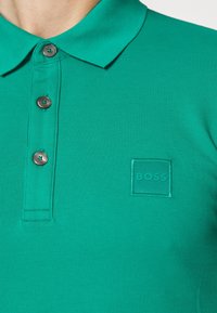 Polo vert en tissu coton texturé avec un col, trois boutons et un patch logo carré avec "BOSS" dans une couleur assortie.