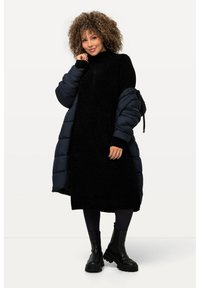 Abito nero in maglia testurizzata con colletto alto. Abbinato a un cappotto imbottito blu navy. La modella indossa leggings scuri e stivali neri chunky.