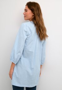 Tunic di colore azzurro chiaro, tessuto traspirante, maniche lunghe con polsini elasticizzati, retro leggermente più lungo e una texture liscia senza motivi.