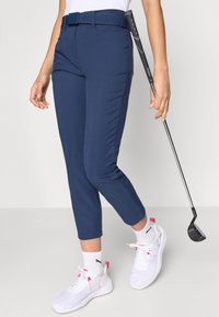 Παντελόνια γκολφ σε ναυτικό μπλε με εφαρμοστή γραμμή και με υφασμάτινη ζώνη με υφή. Συνδυάζονται με λευκά αθλητικά παπούτσια και κρατούν ένα ξύλο γκολφ.