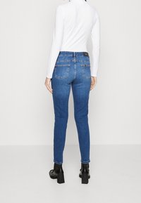 Mujer de espaldas a la cámara, vestida con jeans skinny azules, una camiseta blanca de manga larga y botines negros de tacón en un fondo uniforme.