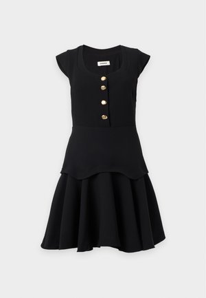 Femme debout portant une robe noire sans manches avec des boutons dorés et une jupe évasée, associée à des chaussures plates noires.