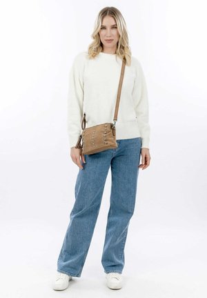 Crossbody bag in tan suede with silver studs. Pairing with a white sweater and blue jeans, model stands against a white background.