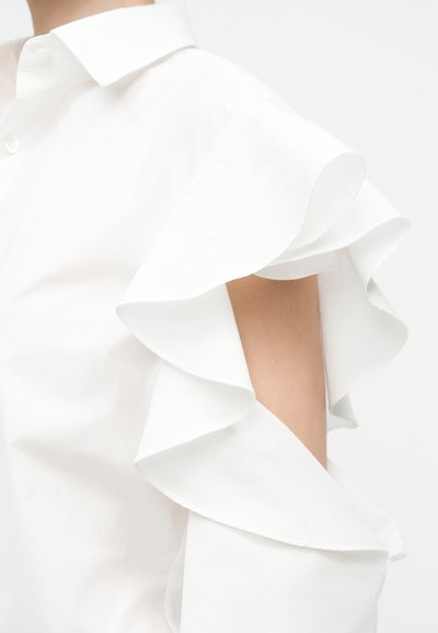 Bluza albă cu detaliu stratificat cu volane pe umărul descoperit care arată brațul superior gol, cu guler cu nasturi vizibil.