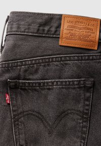 Jeans en denim noir avec une étiquette en cuir marron à la taille, présentant des coutures courbes sur la poche arrière et une petite étiquette rouge Levi's.