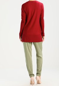 Długi czerwony dziany sweter o prążkowanej fakturze połączony z jasnozielonymi spodniami zwężanymi i beżowymi szpilkami, prezentujący swobodną sylwetkę.