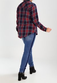 Rood- en marineblauwe geruite blouse met lange mouwen en een relaxte pasvorm. Draagt bijpassende donkerblauwe denim jeans met een zijstreep. Zwarte enkellaarsjes met hak.