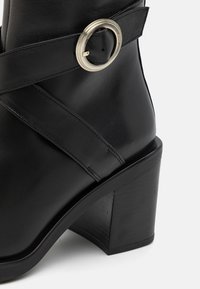 Czarny skórzany botek za kostkę z zaokrąglonym noskiem, masywnym obcasem i ozdobnym srebrnym klamrą na krzyżującym się pasku. Gładka tekstura.
