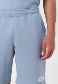 Pantaloni corti di cotone blu chiaro con un elastico in vita, tasche laterali e un logo bianco sulla parte inferiore della gamba sinistra. Texture liscia, design casual.