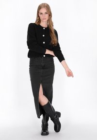 Schwarze gerippte Strickjacke mit drei großen weißen Knöpfen, kombiniert mit einem schwarz-hochgeschnittenen Denimrock und kniehohen schwarzen Stiefeln.